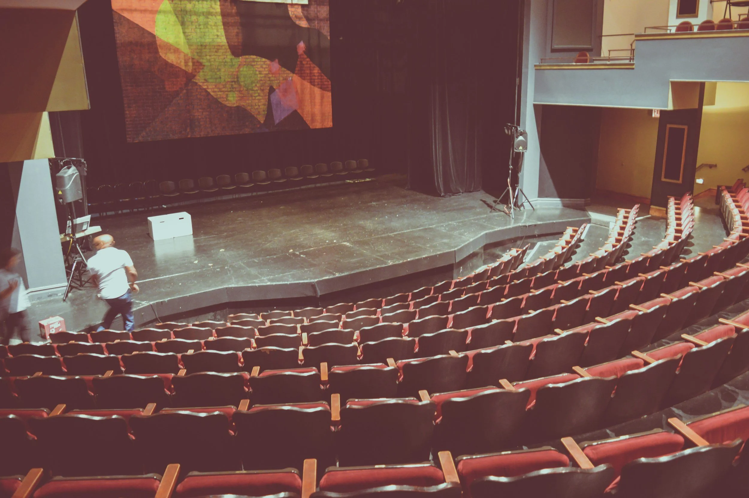 An empty theater with red seats, a stage with black curtains, a large colorful abstract artwork projected on the back wall, and a few people setting up equipment.