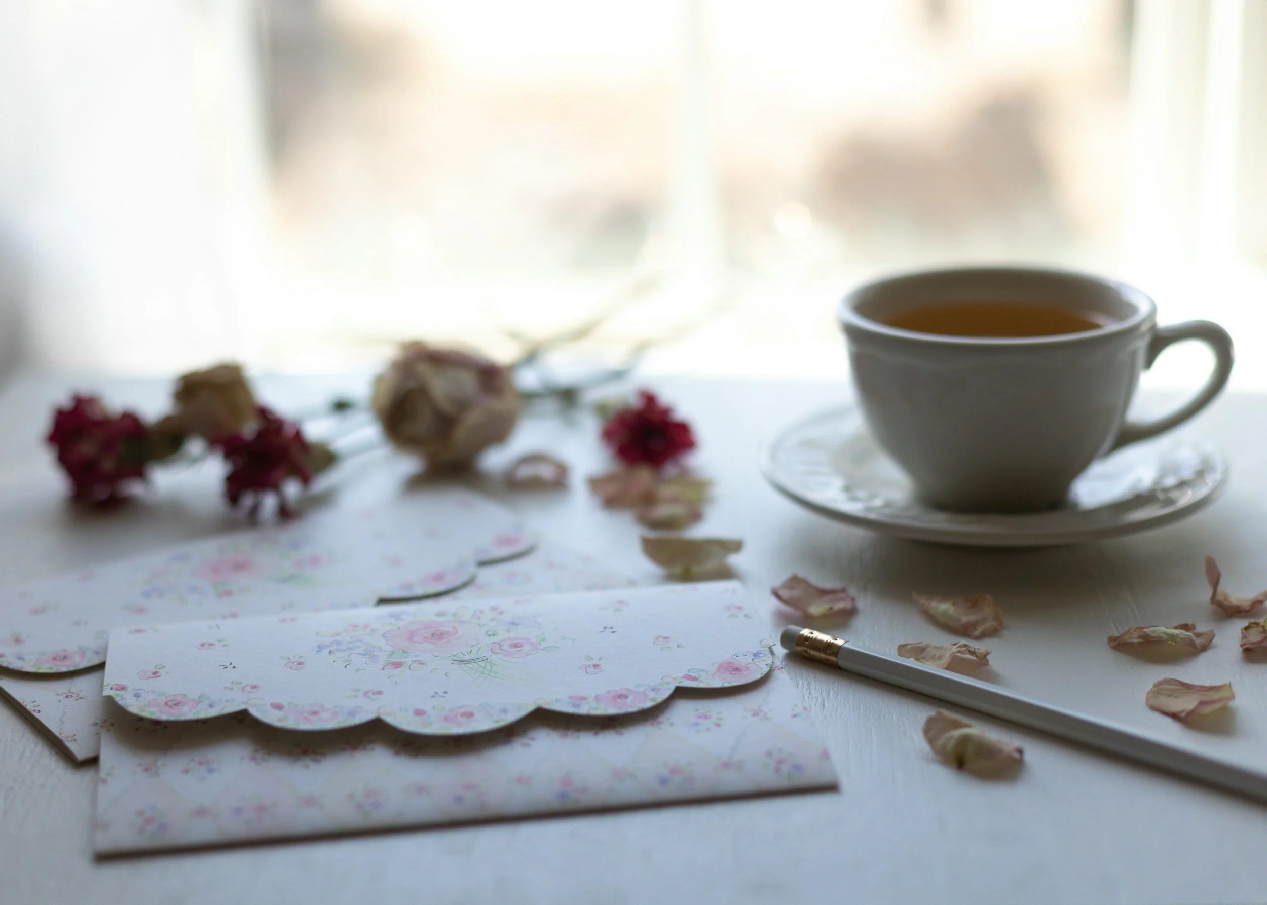 A white table with scattered floral-patterned papers, a gold and white pen, dried flower petals, a white cup of tea on a saucer, and some blurred pink flowers in the background near a window with light streaming through.