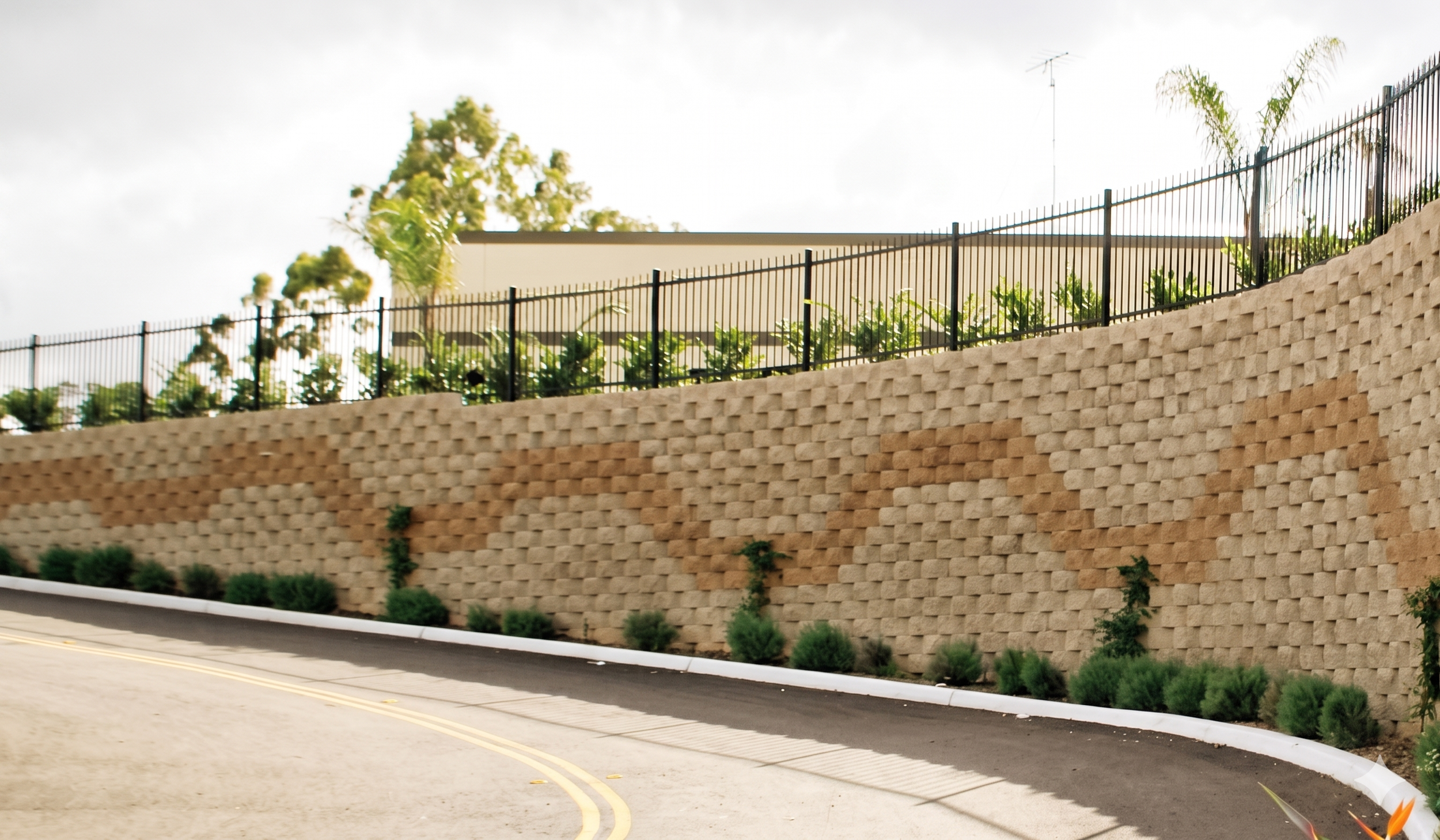 Muro de bloques con vegetación y un lecho de cubo de piedra decorativa, con una cerca de metal en la parte superior, junto a una calle asfaltada con línea amarilla central y pequeños arbustos en la acera.