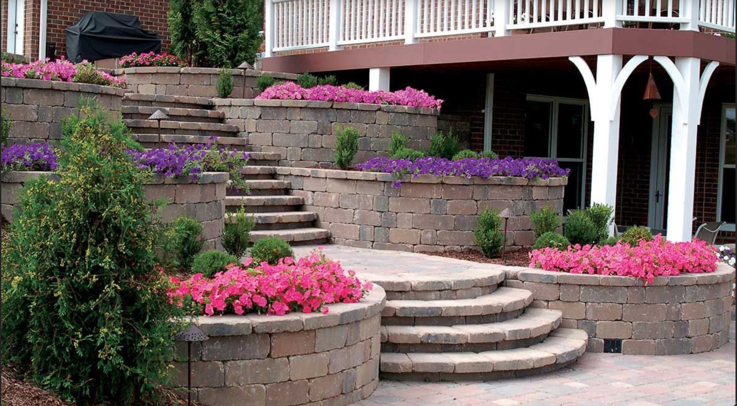 Escalera de piedra que conecta dos niveles de un jardín con muchas flores rosas y moradas, junto a un porche con barandillas blancas.