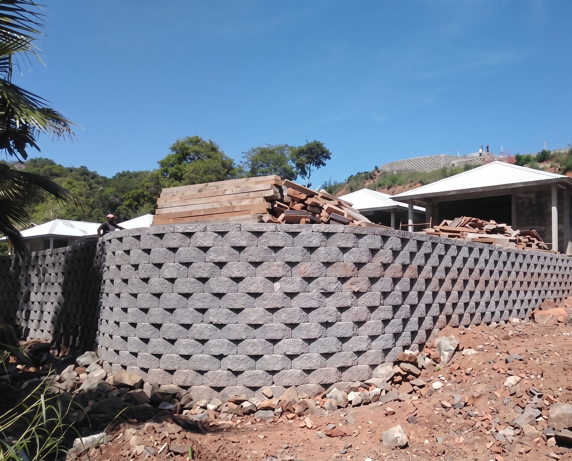 Muro de bloques de concreto en construcción con madera apilada en la parte superior y edificios con techos blancos en el fondo, en un área rural bajo un cielo despejado.