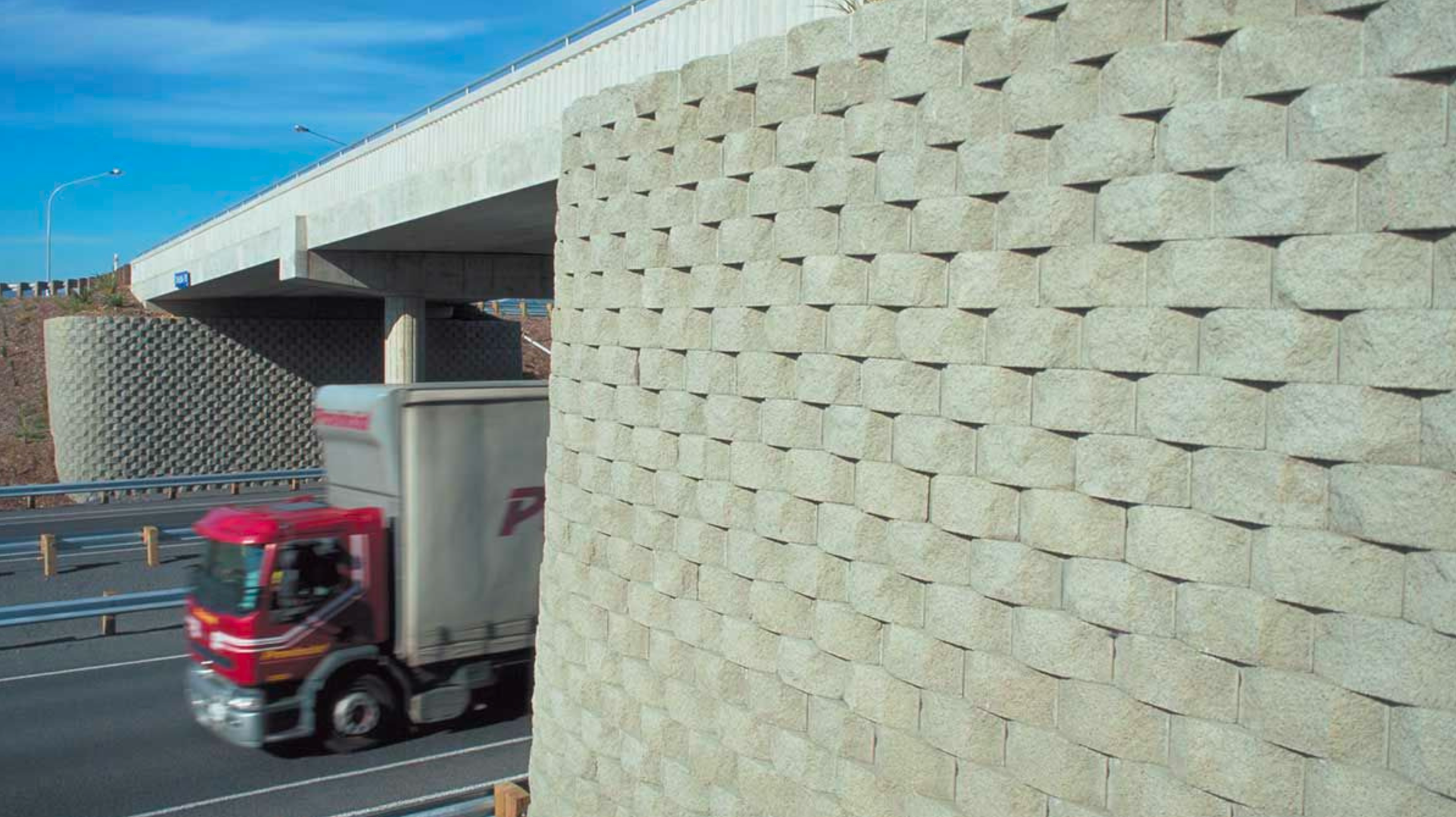 Vista de un puente elevado de concreto con un muro de bloques de piedra en primer plano y un camión de carga en movimiento en la autopista debajo del puente, con cielo despejado.