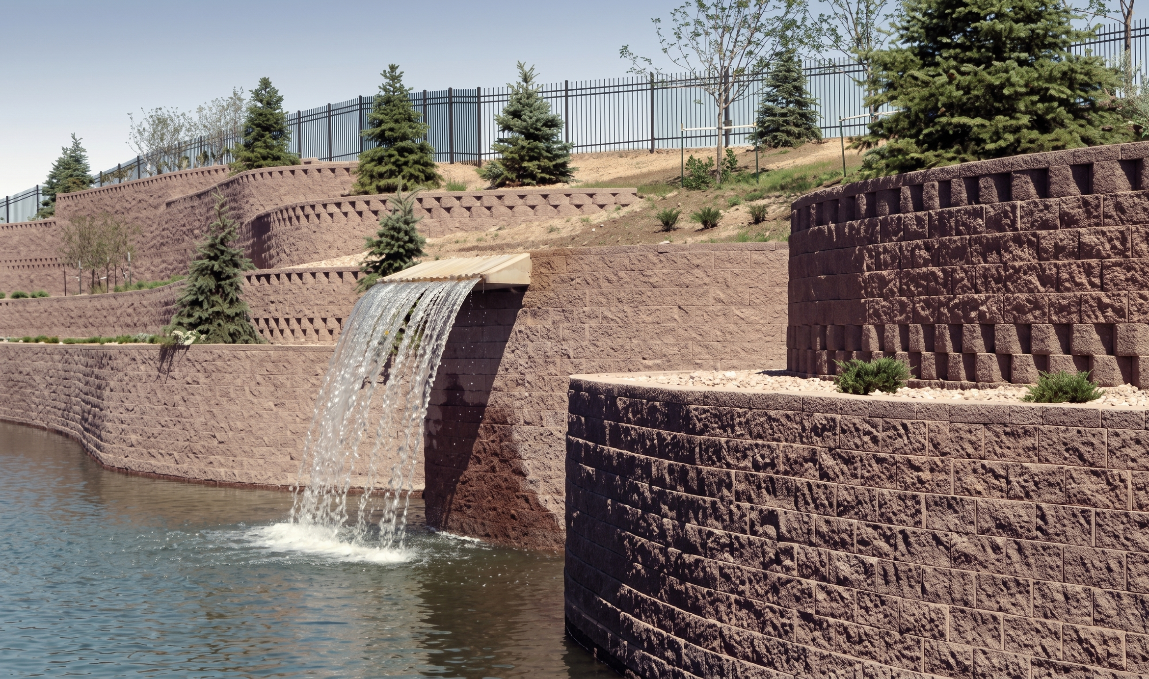 Muros de piedra decorativos en terraplén con árboles y fuente de agua en un parque.