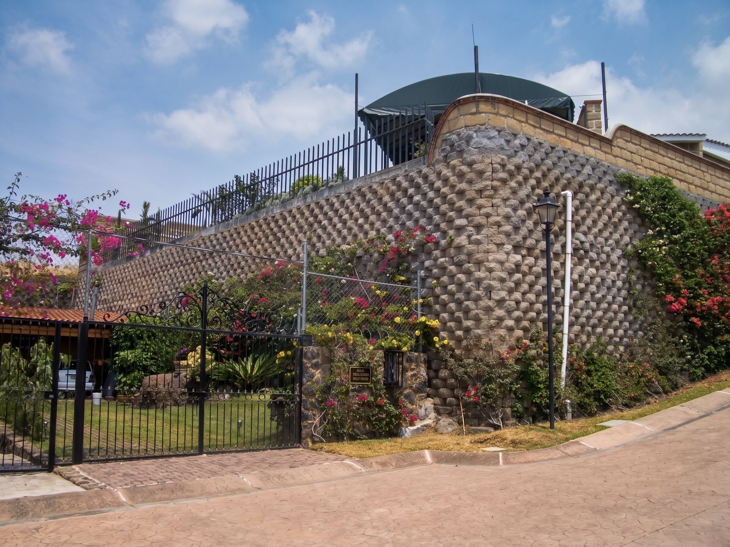 Entrada de la Villa Esmeralda con reja negra, área ajardinada con flores coloridas y señal en la puerta.