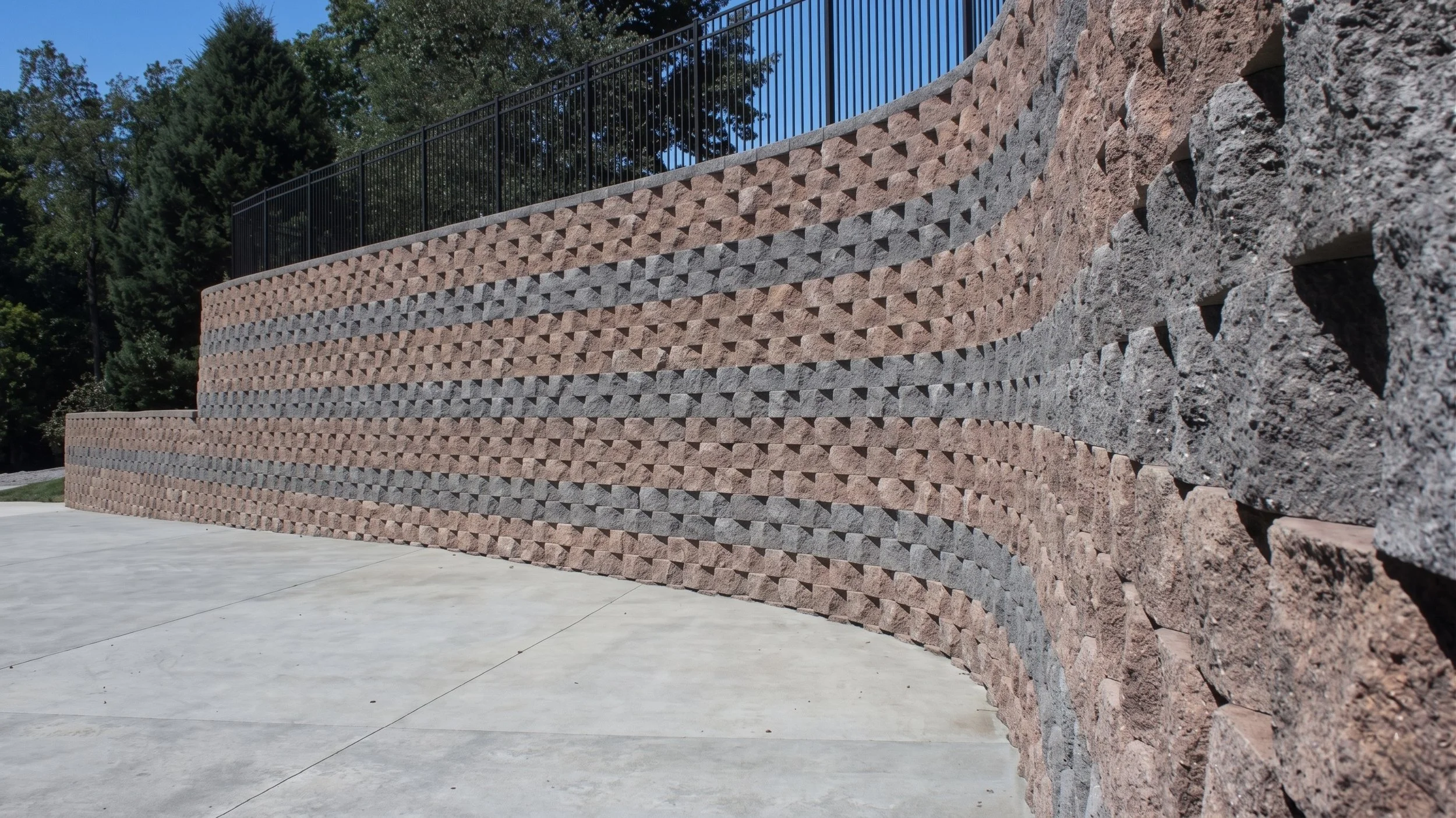 Muro de bloques de piedra decorativos en tonos rojos y grises, con una cerca metálica negra en la parte superior, junto a árboles verdes y un cielo azul.