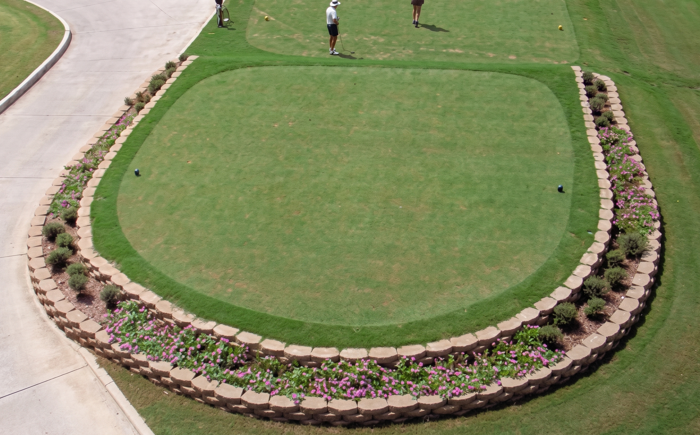 Vista aérea de un campo de golf con césped, rodeado por una pared de piedras y plantas, y personas jugando al golf.