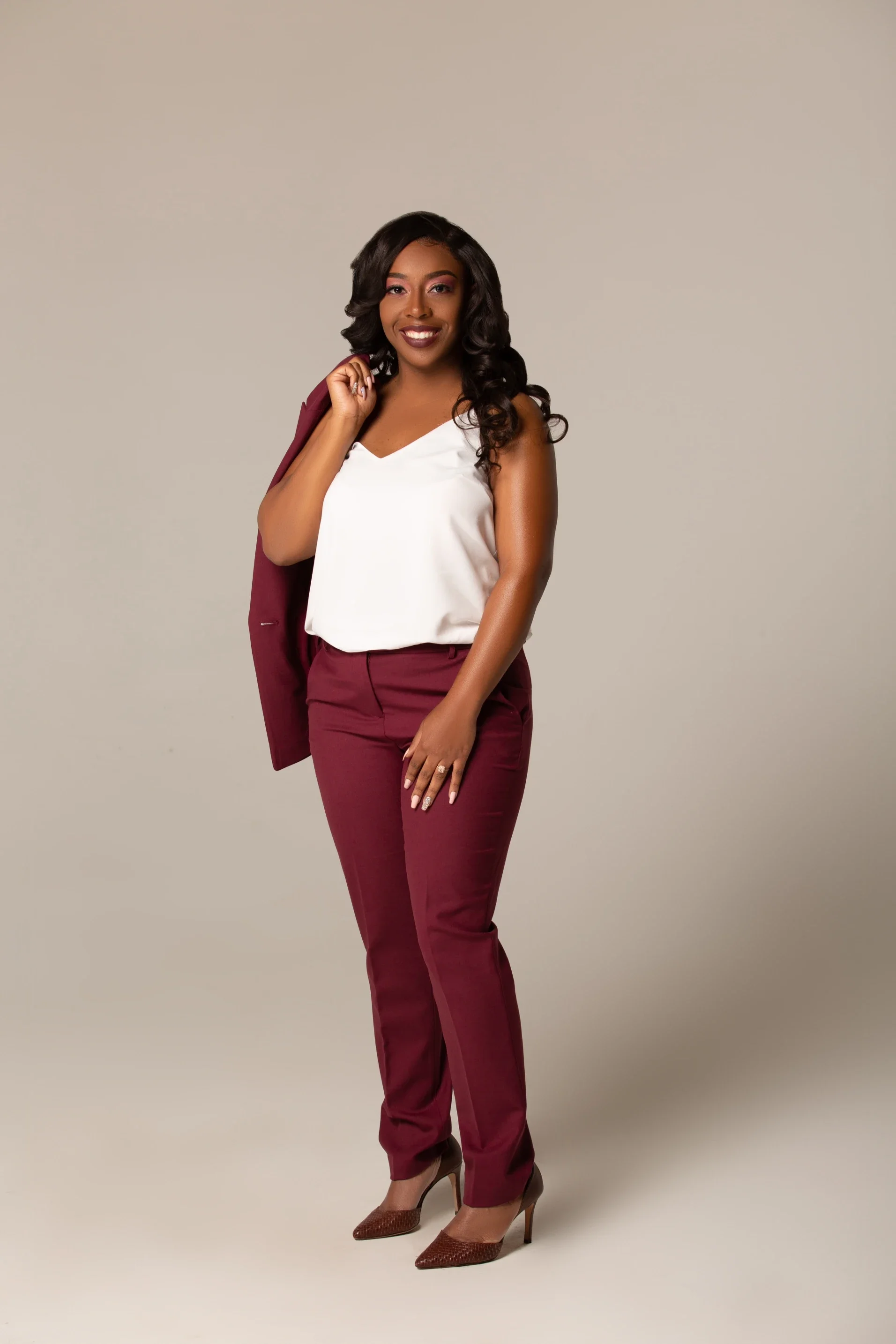 A woman with dark hair styled in loose curls stands against a neutral background, wearing a white sleeveless top, burgundy suit pants, a matching blazer over her shoulder, and high-heeled shoes, smiling confidently.