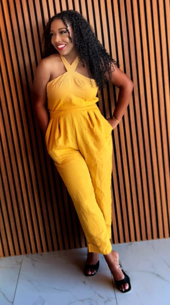 A woman in a yellow jumpsuit with halter straps, standing against a wooden slatted wall, smiling with black curly hair, wearing black high heels.