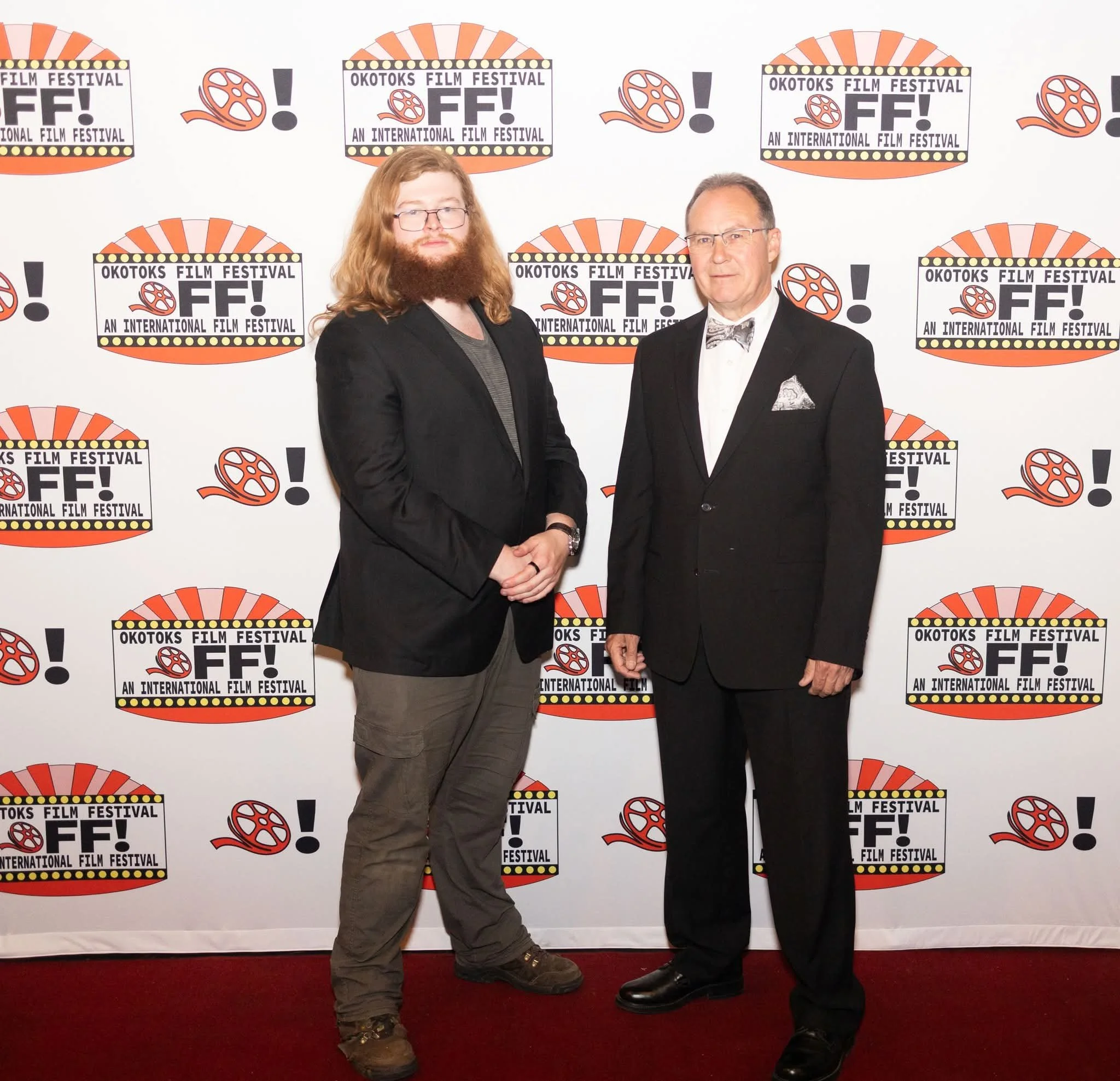 Two men in tuxedos standing in front of a step and repeat banner that reads "Okotoks Film Festival - An International Film Festival" with film reel and exclamation mark graphics.