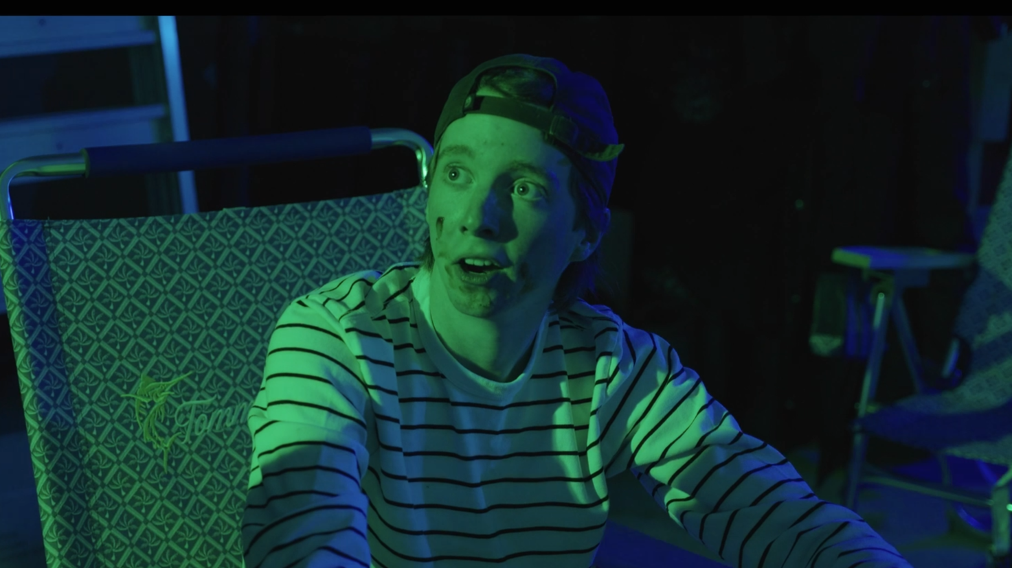 Person with striped shirt and backwards cap sitting outdoors at night, illuminated by green and blue lights, with a camping chair behind.