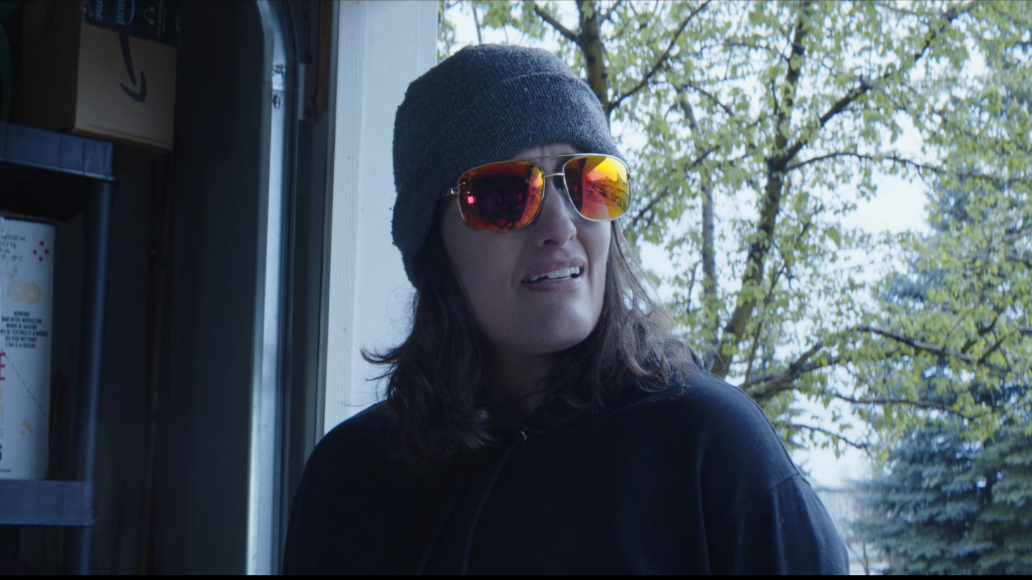 A woman wearing sunglasses, a grey beanie, and a black hoodie looks confused or displeased outside near trees.