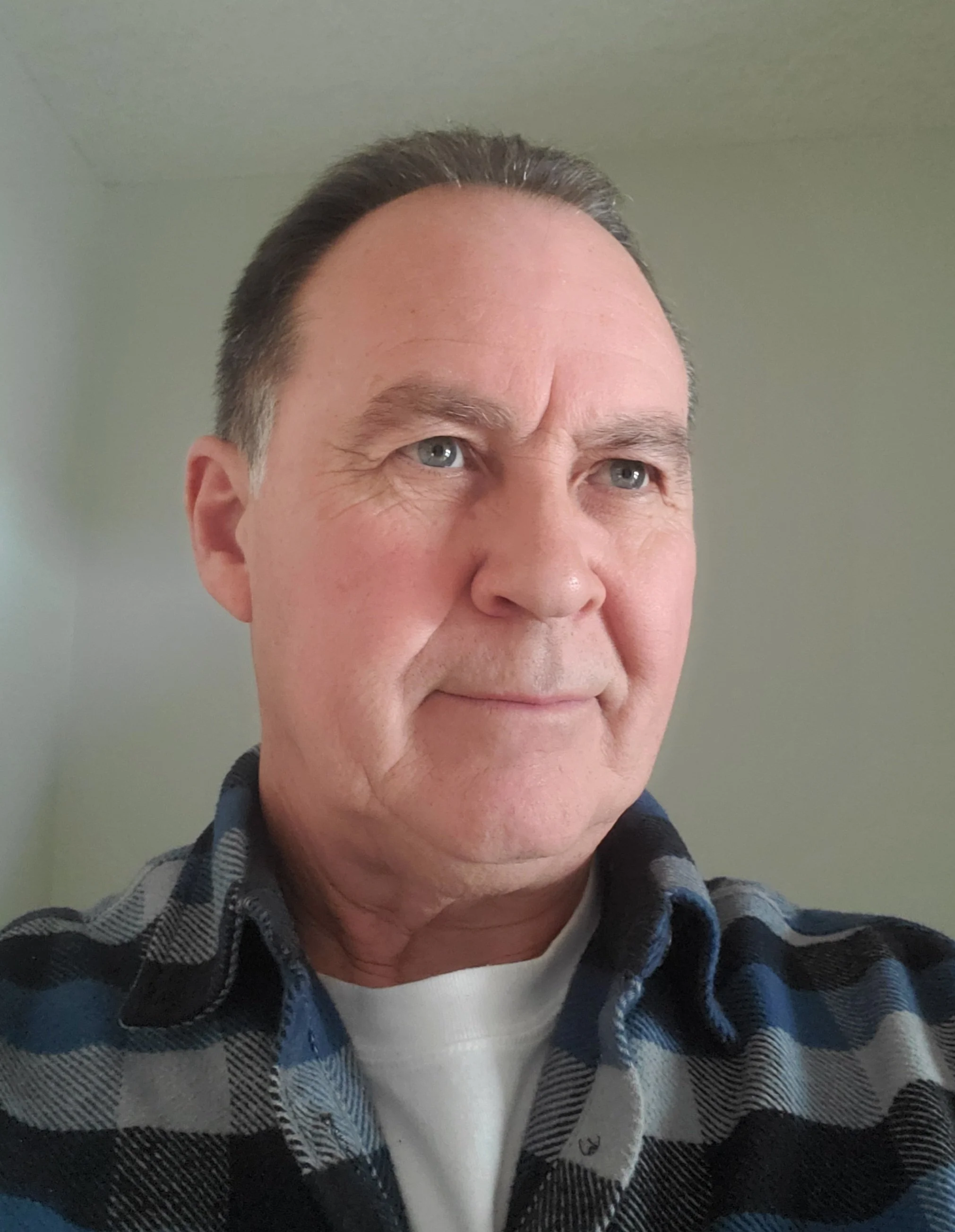 Close-up of a middle-aged man's face, showing his forehead, eyes, and part of his nose and mouth. He has short dark hair and is wearing a plaid shirt over a white T-shirt, with a neutral background.