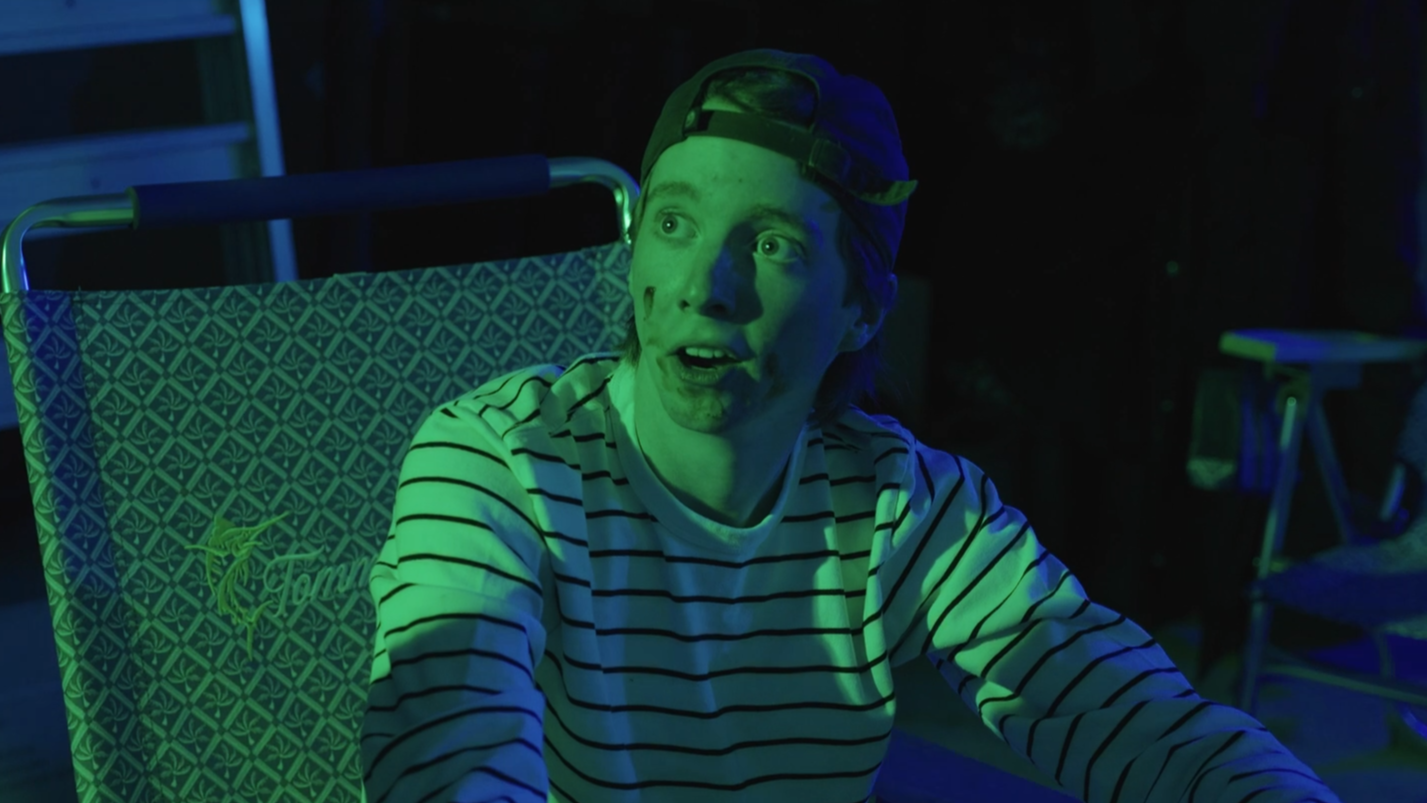 A young man with a striped shirt and backwards cap sitting outdoors at night, illuminated by colorful lights, with an expressive surprised or shocked facial expression.
