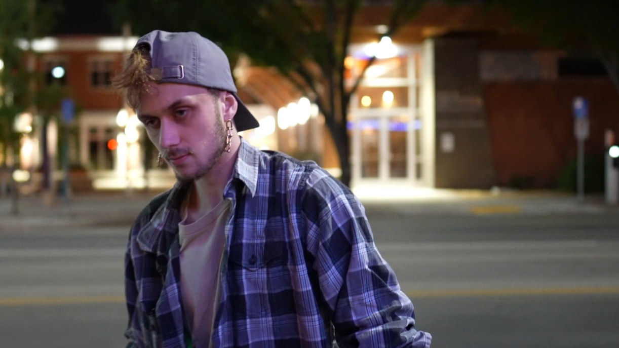Young man with light brown hair wearing a backwards gray cap, purple and black plaid shirt, and earrings, standing on a city street at night with blurry lights and buildings in the background.