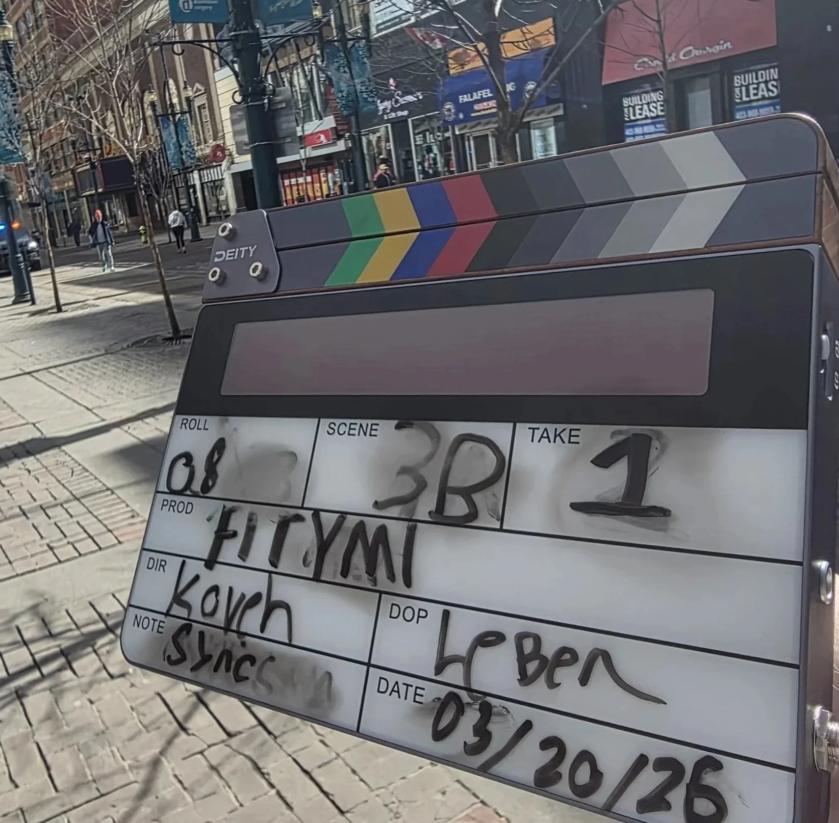 A city sidewalk with shops and pedestrians, and a clapperboard for filmmaking positioned in the foreground.