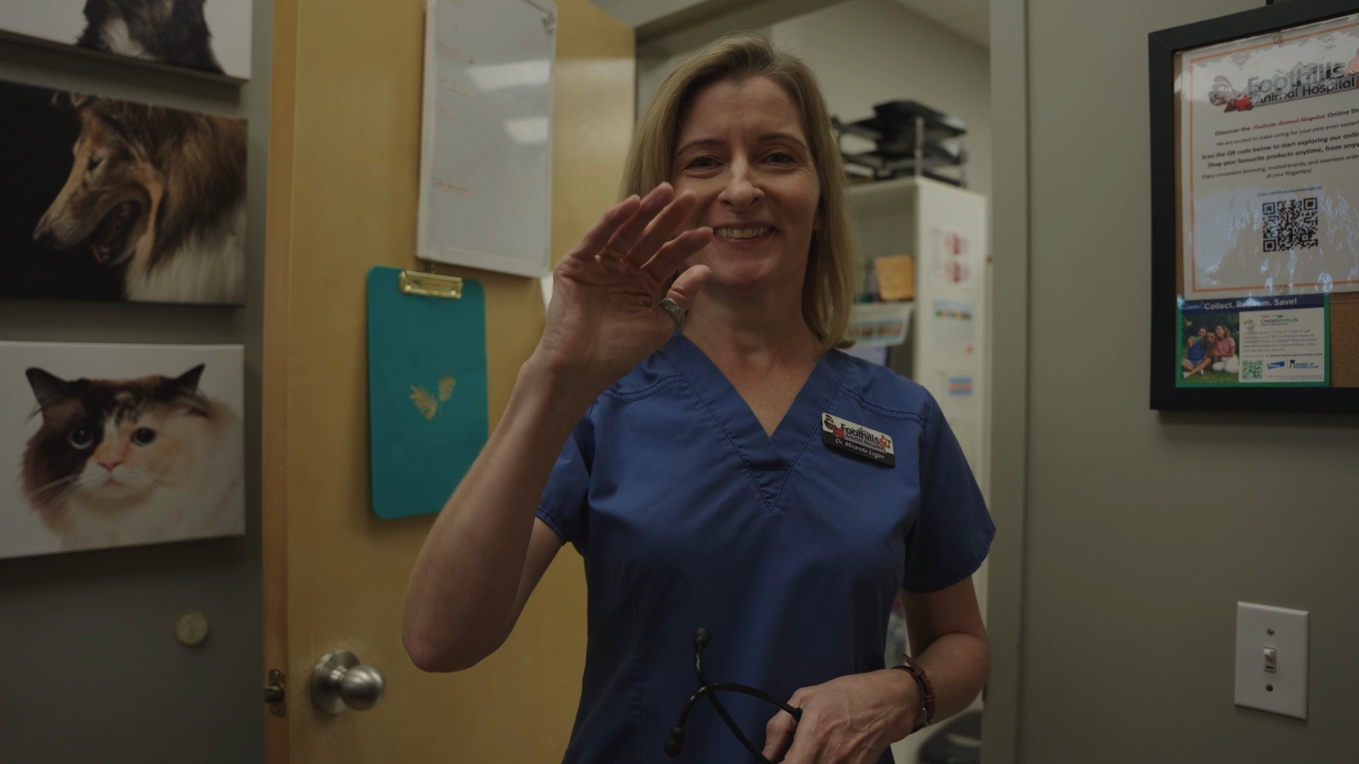 A veterinarian in blue scrubs smiling and waving inside an animal clinic, with pictures of a dog and a cat on the wall behind her.