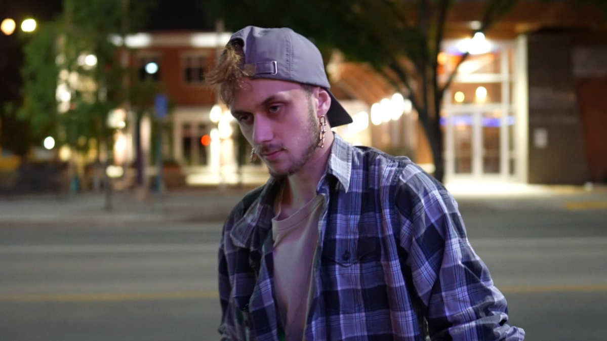Young man with a light beard and wearing a gray cap backwards, plaid shirt, and earrings, standing on a city street at night with blurred buildings and lights in the background.