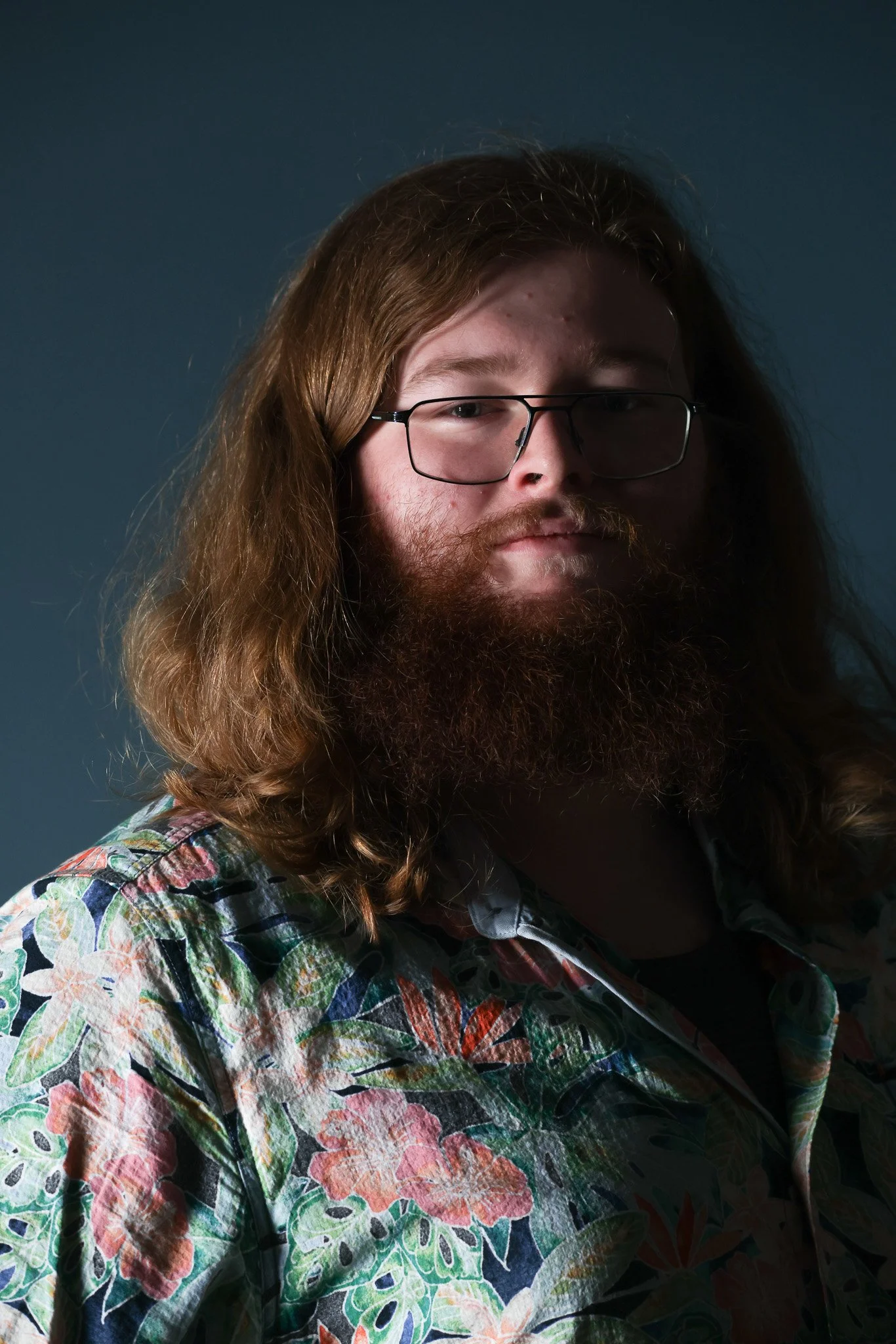 Close-up of a young man with long hair, beard, glasses, wearing a floral shirt, against a dark background.