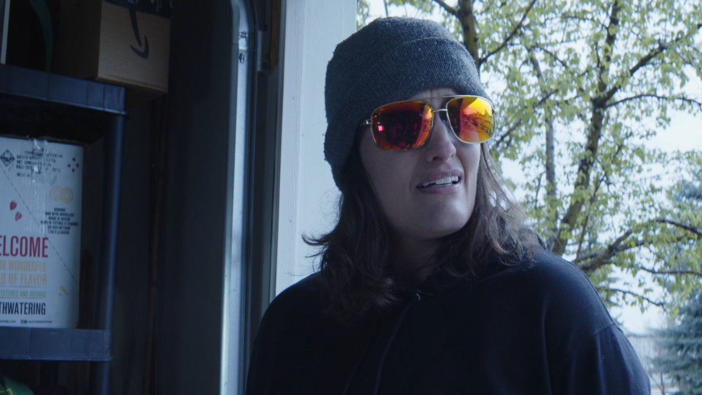 A woman wearing a gray beanie, sunglasses with reflective lenses, and a black hoodie, looking to her right outside a window with trees in the background.