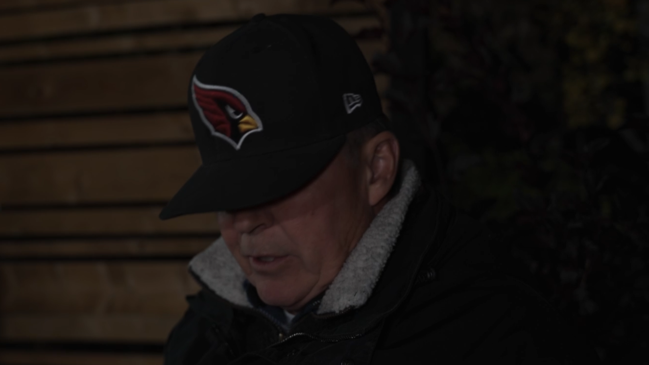 An older man wearing a black hat with a sports team logo, partially obscuring his face, outdoors at night.