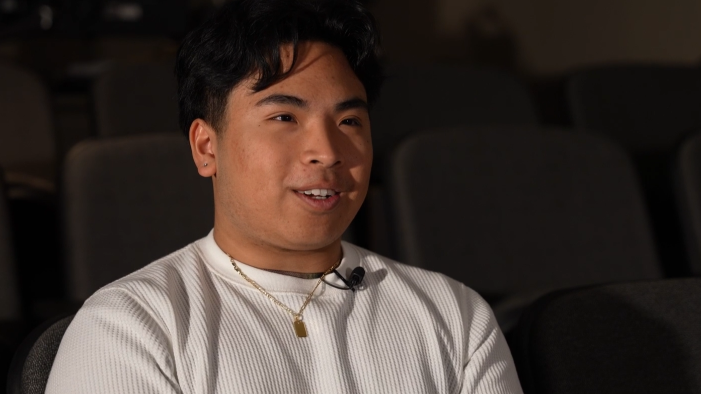 A young man sitting in an auditorium with black chairs, wearing a white shirt, a gold necklace, and a small black microphone clipped to his shirt.