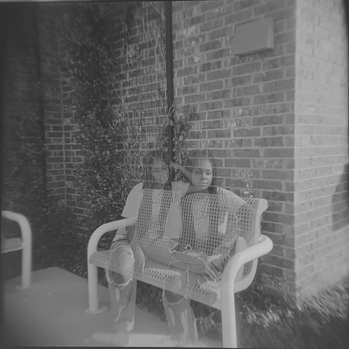 A black and white photo of a young girl sitting on a bench outside against a brick wall, with a reflection of her face and arm visible on the glass window in front of her.