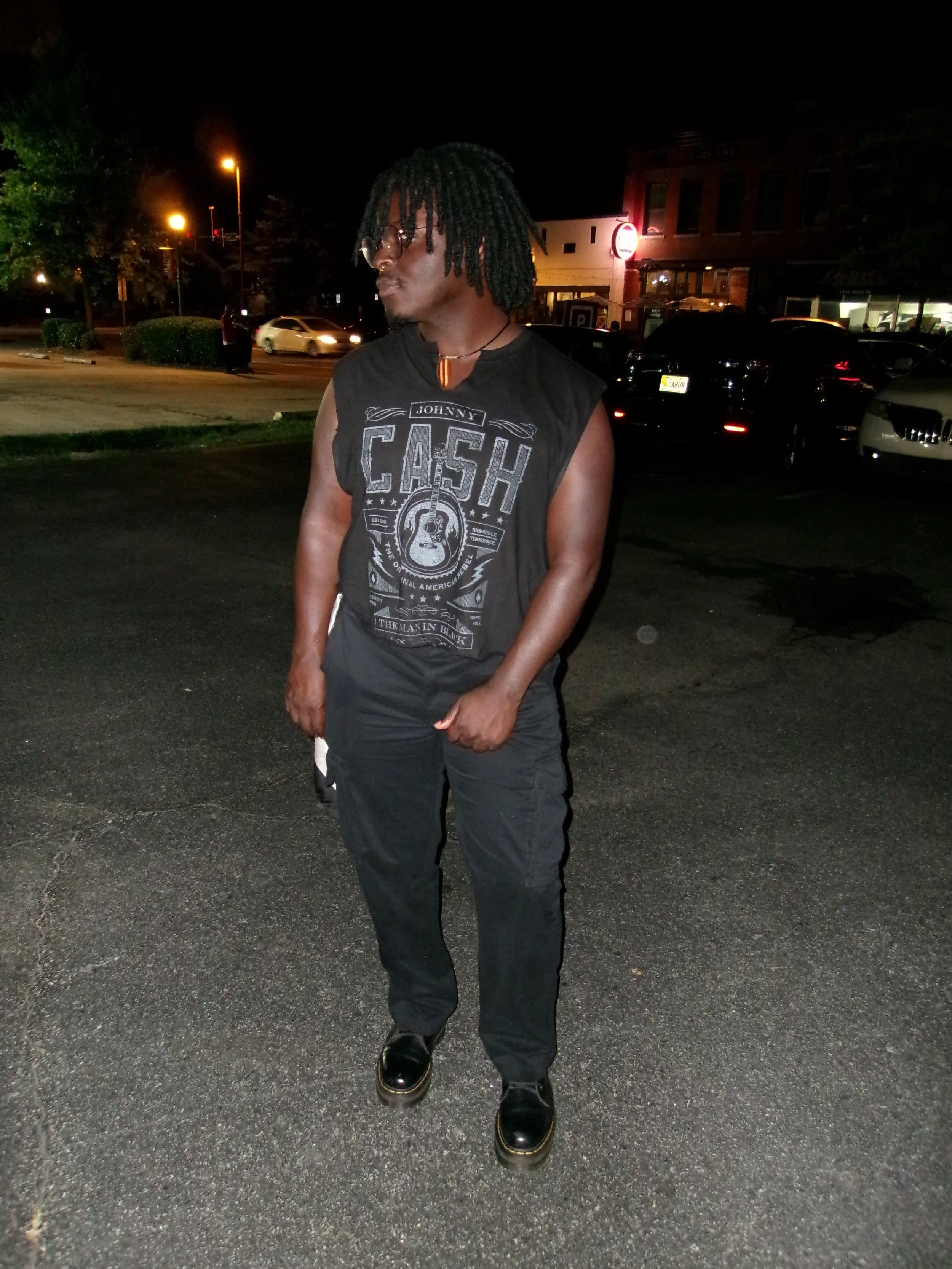 A person with dreadlocks wearing glasses, a sleeveless black graphic shirt, black pants, and black shoes standing on a dark parking lot at night, with cars and illuminated buildings behind him.