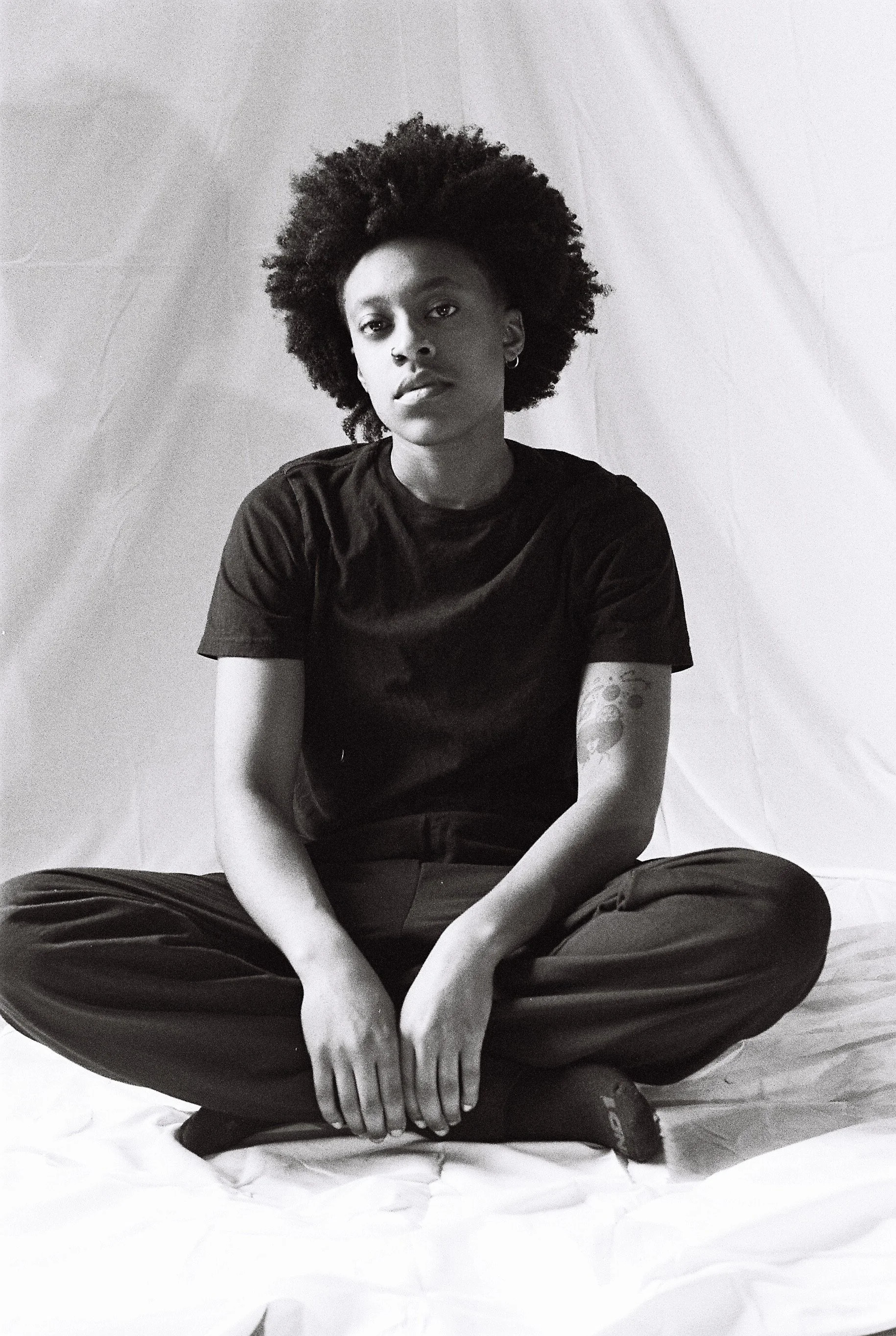 Black and white photo of a woman sitting cross-legged on a white surface, looking directly at the camera with a neutral expression, wearing a dark T-shirt and dark pants, and having a tattoo on her left arm.