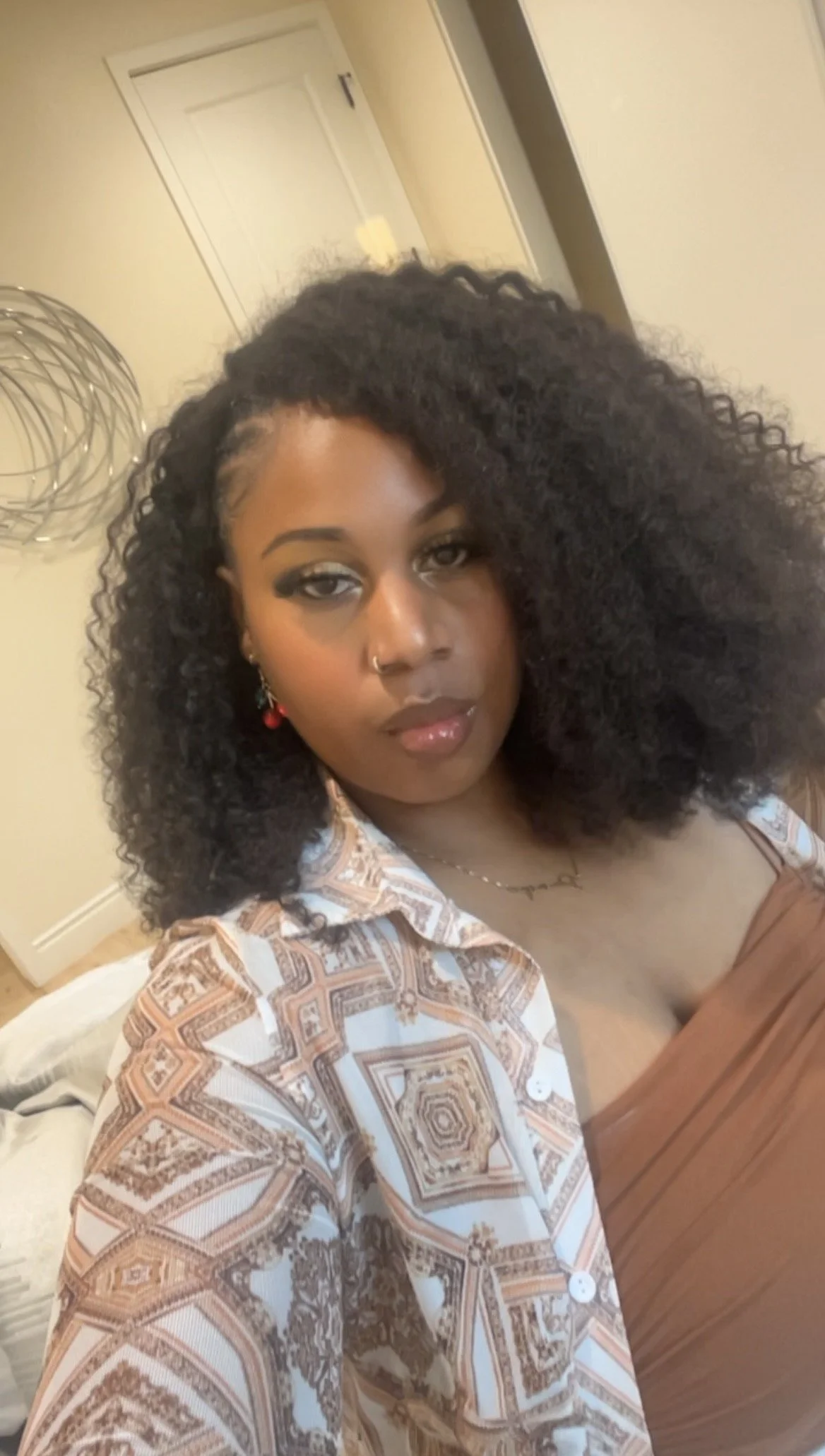 A woman with curly black hair, makeup, and earrings standing indoors in front of a mirror.
