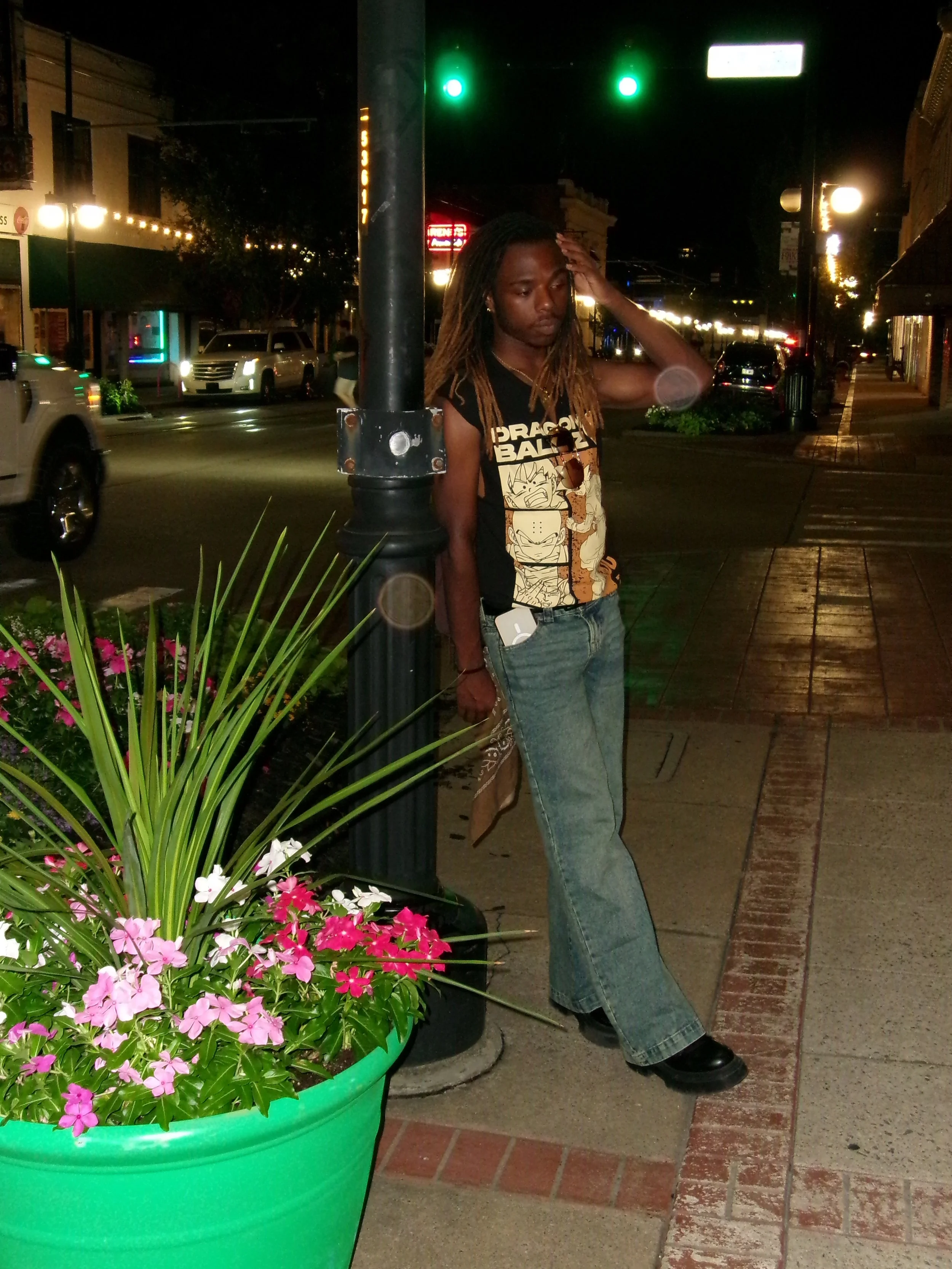 A woman with dreadlocks and wearing a Dragon Ball Z t-shirt is standing on a city sidewalk at night, leaning against a lamppost, with her hand on her forehead. There are cars on the street and illuminated storefronts in the background. A large green flowerpot with pink and white flowers is in the foreground.