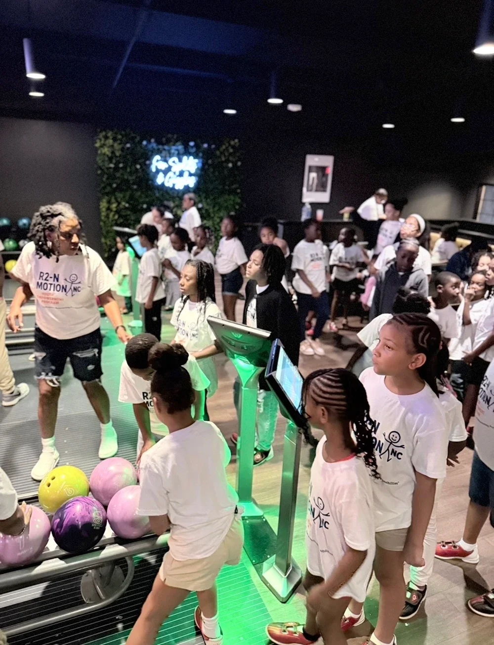 Children at a bowling alley with colorful bowling balls in the foreground, many children wearing white T-shirts, and an adult instructor guiding them. The background shows a dark room with a neon sign and arcade games.