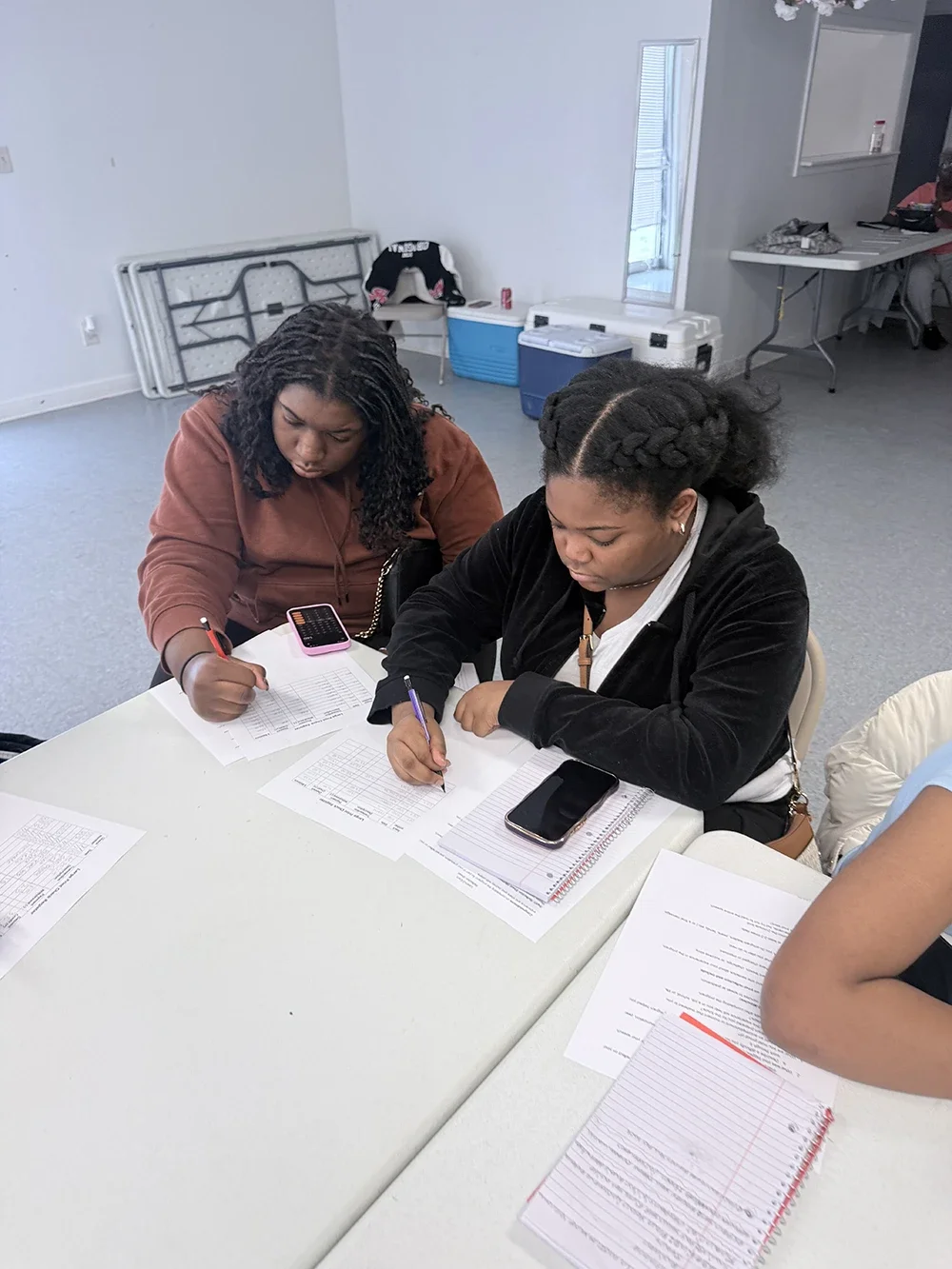 Two women sitting at a white table, working on documents with pens, a calculator, and a smartphone. There are additional papers and notebooks on the table, in a room with a white wall, window, and some folded furniture or equipment against the wall.