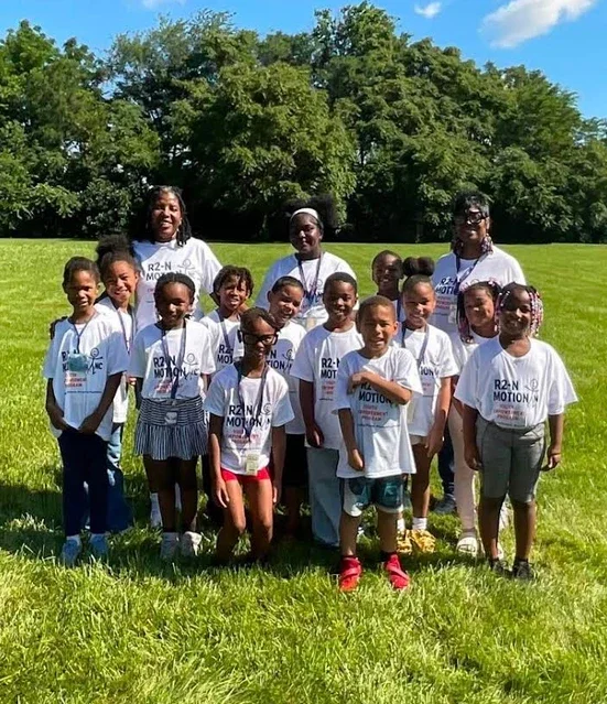 Group of children and two adults standing outdoors in a grassy field on a sunny day, all wearing white T-shirts with 'R2N Motion' printed on them.