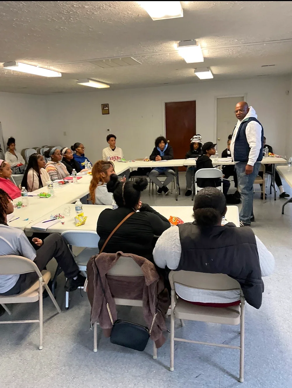 A guide is speaking to a diverse group of people seated around a U-shaped arrangement of tables in a room with white walls and fluorescent ceiling lights.