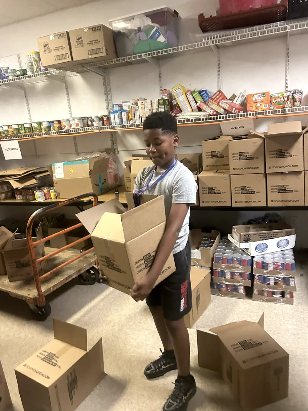 A boy organizing boxes in a storage room with shelves filled with canned goods, boxed food, and other supplies.