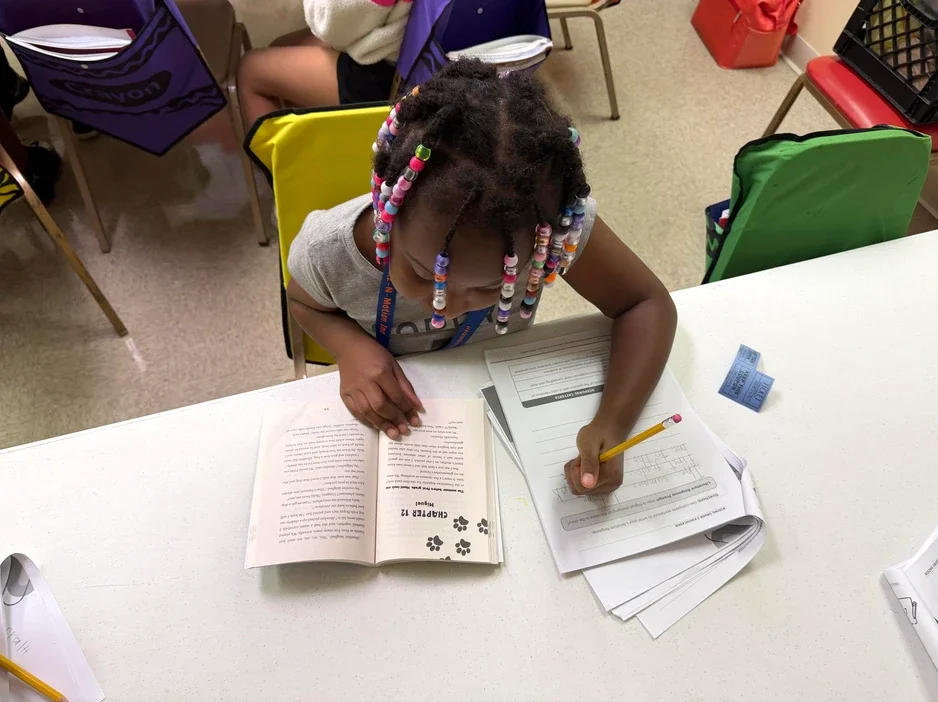 A young girl with colorful beaded hair sitting at a desk, studying with a book and worksheets in a classroom.