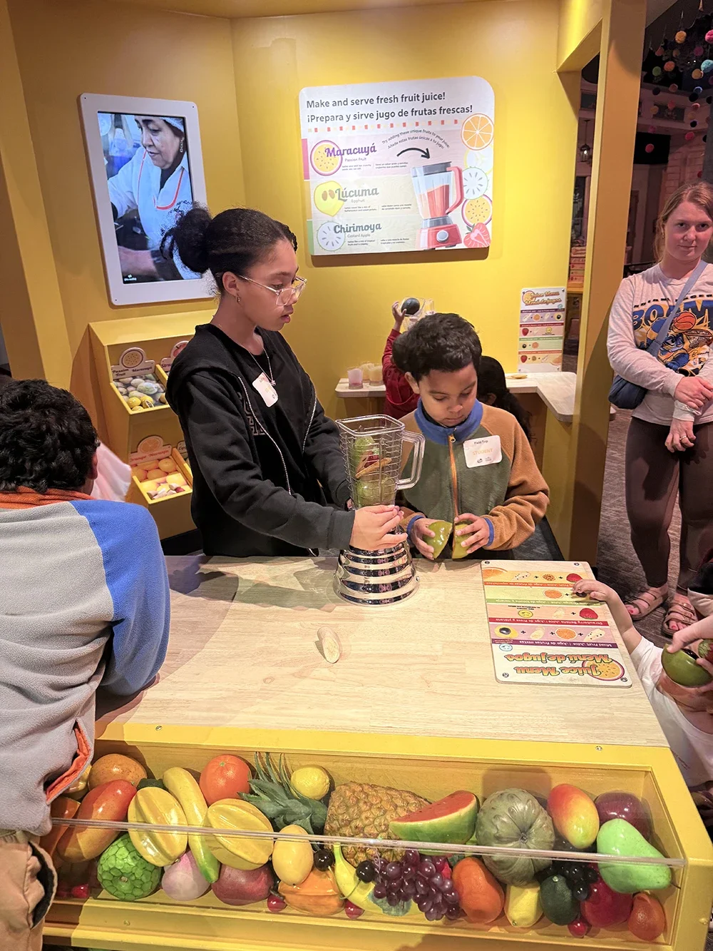 Children participating in a fruit juice-making activity at a display table with various fruits, a blender, and informational posters on the yellow wall behind them.