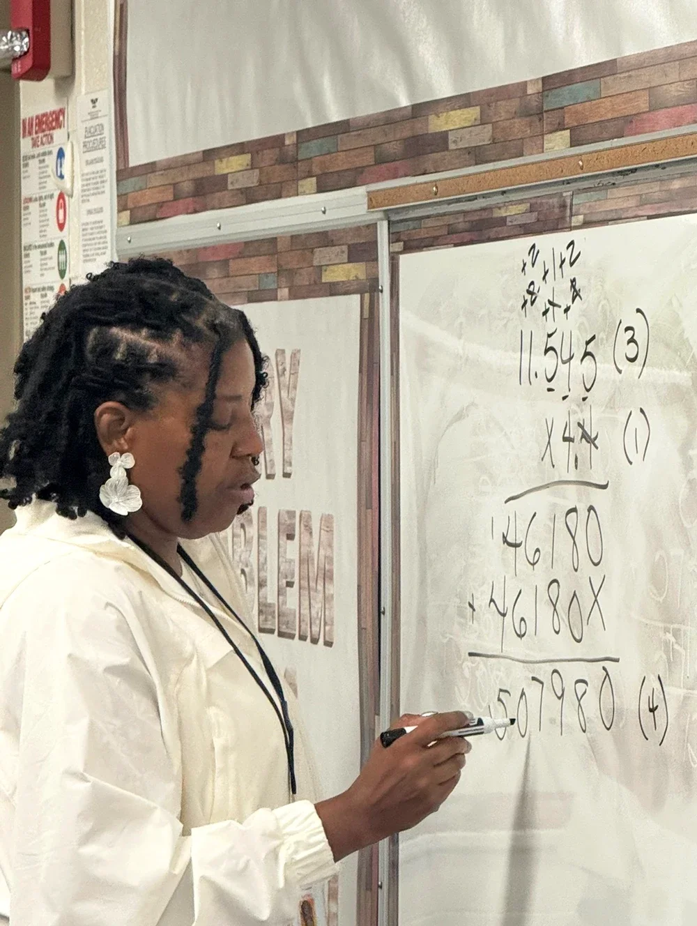 A woman with dreadlocks and butterfly earrings writes on a whiteboard in a classroom.