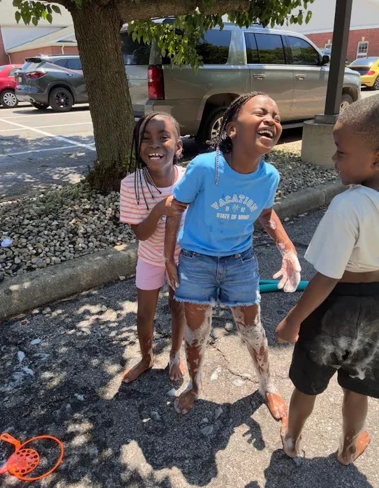 Three children playing outside on a sunny day, with two girls laughing and a boy watching them, covered in soap suds.