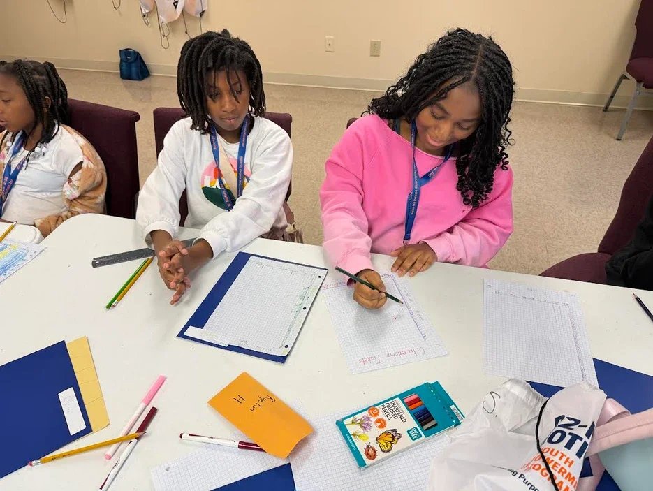 Three young girls sitting at a table in a classroom, working on school assignments with notebooks, pencils, and other supplies.