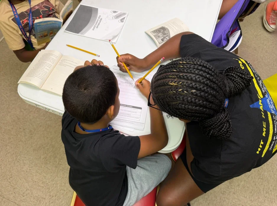 A young boy and a girl with braided hair working together on schoolwork at a table, surrounded by books and supplies.