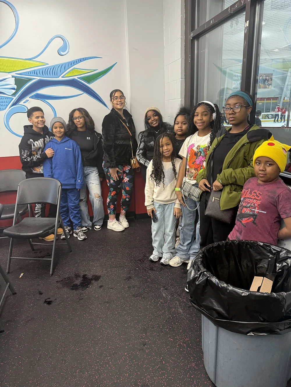 Group of children and two adults standing together indoors near a window, with a colorful mural on the wall behind them and chairs nearby.