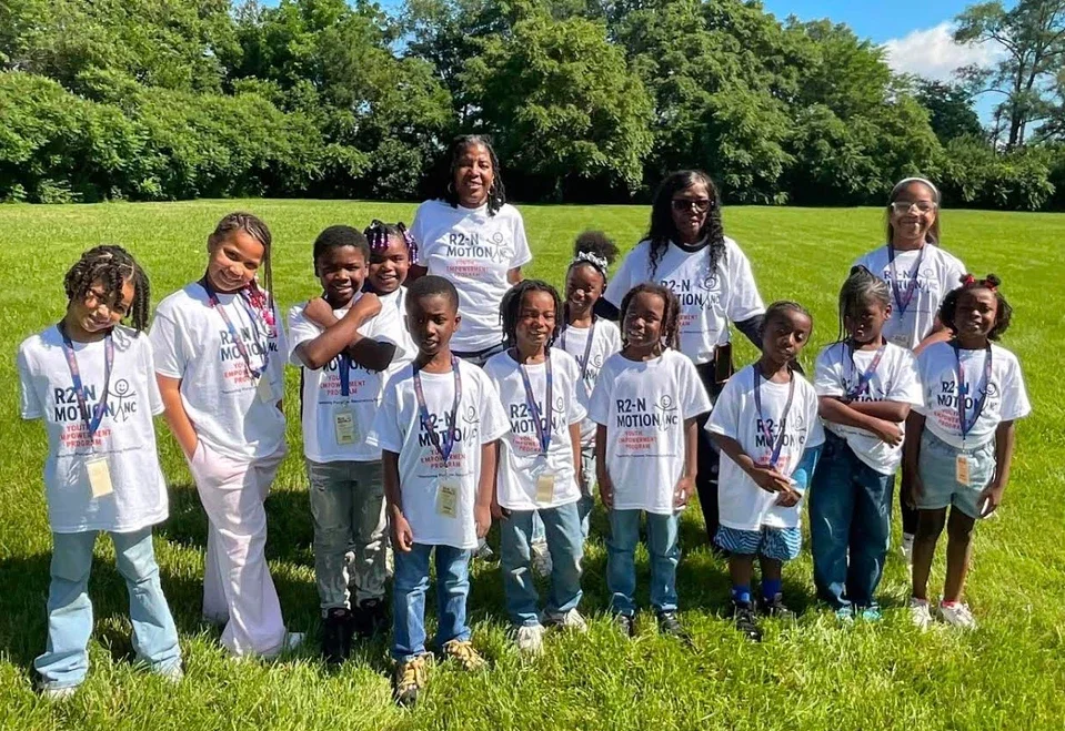 Group of children and adults outdoors on a grassy field, wearing white T-shirts with 'R2N MOTION' printed on them, all smiling for the photo.