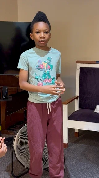 Young girl standing indoors near a chair, wearing a light green T-shirt with colorful characters and maroon pants.