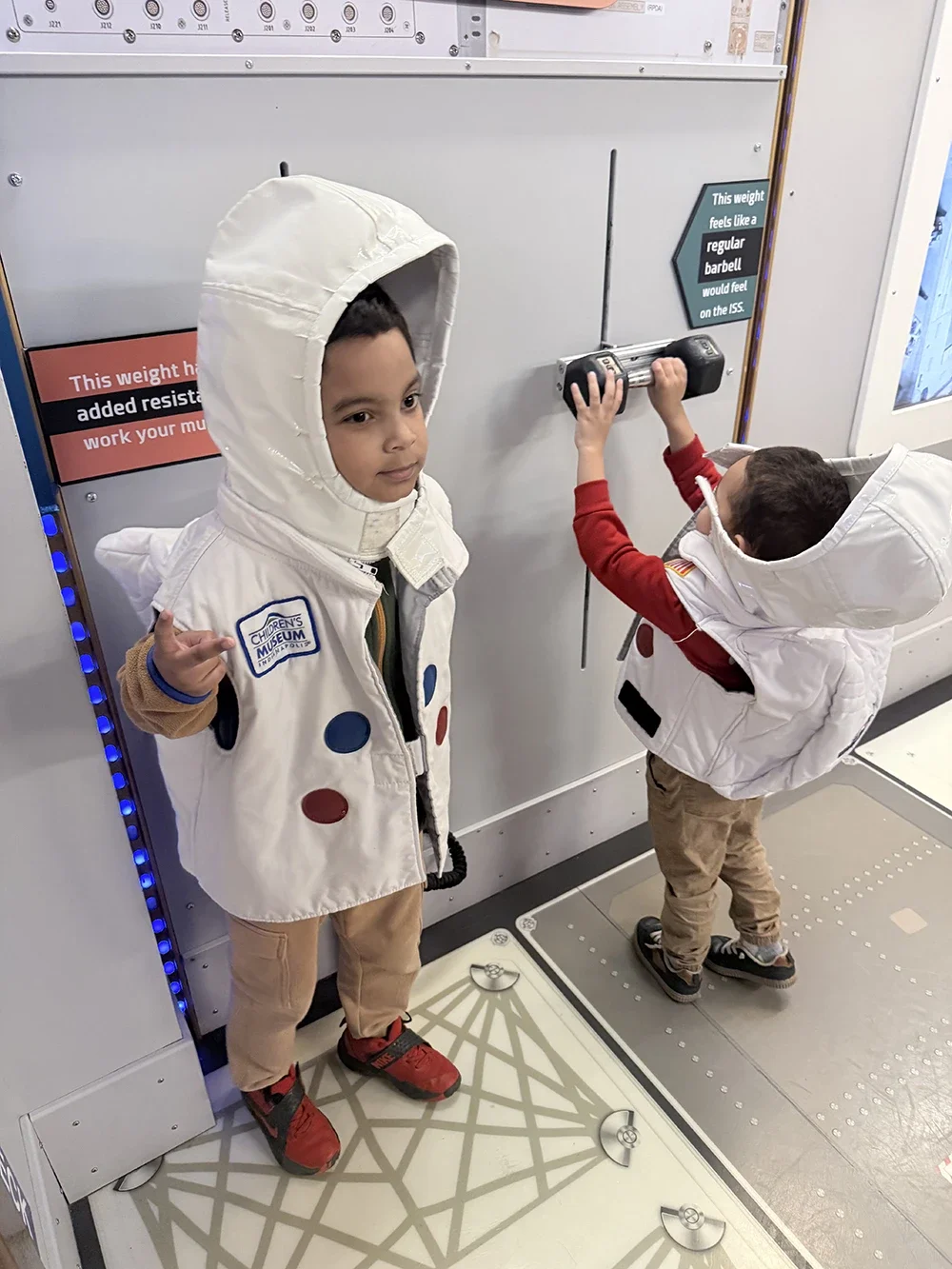 Two children wearing astronaut costumes and hats with hoods, standing in front of a space-themed display with signs about weight and resistance. One child is pointing with his index finger, and the other is lifting a small weight bar.