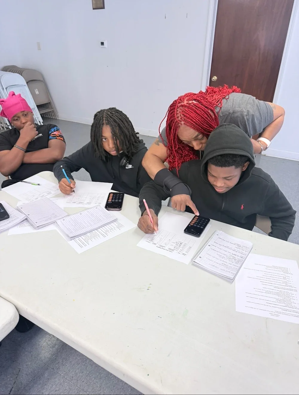 A woman with red braids leans over a table helping three children with academic work. The table is filled with papers, notebooks, and calculators.