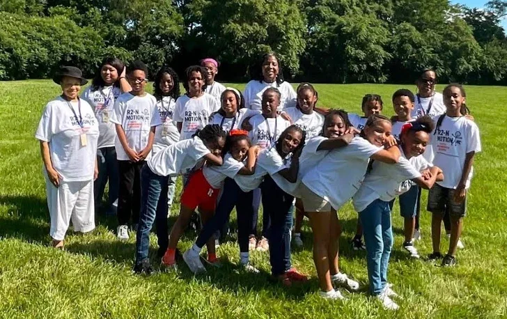 Group of children and adults outdoors on a grassy field, some making a playful train formation, wearing white t-shirts with text, in a sunny park.
