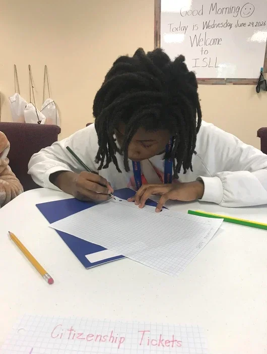 A young person with dreadlocks is writing on a piece of graph paper at a desk, with a yellow pencil nearby. Behind them, a whiteboard displays a welcome message for ISLL, dated June 24, 2026. The setting appears to be a classroom or activity room.