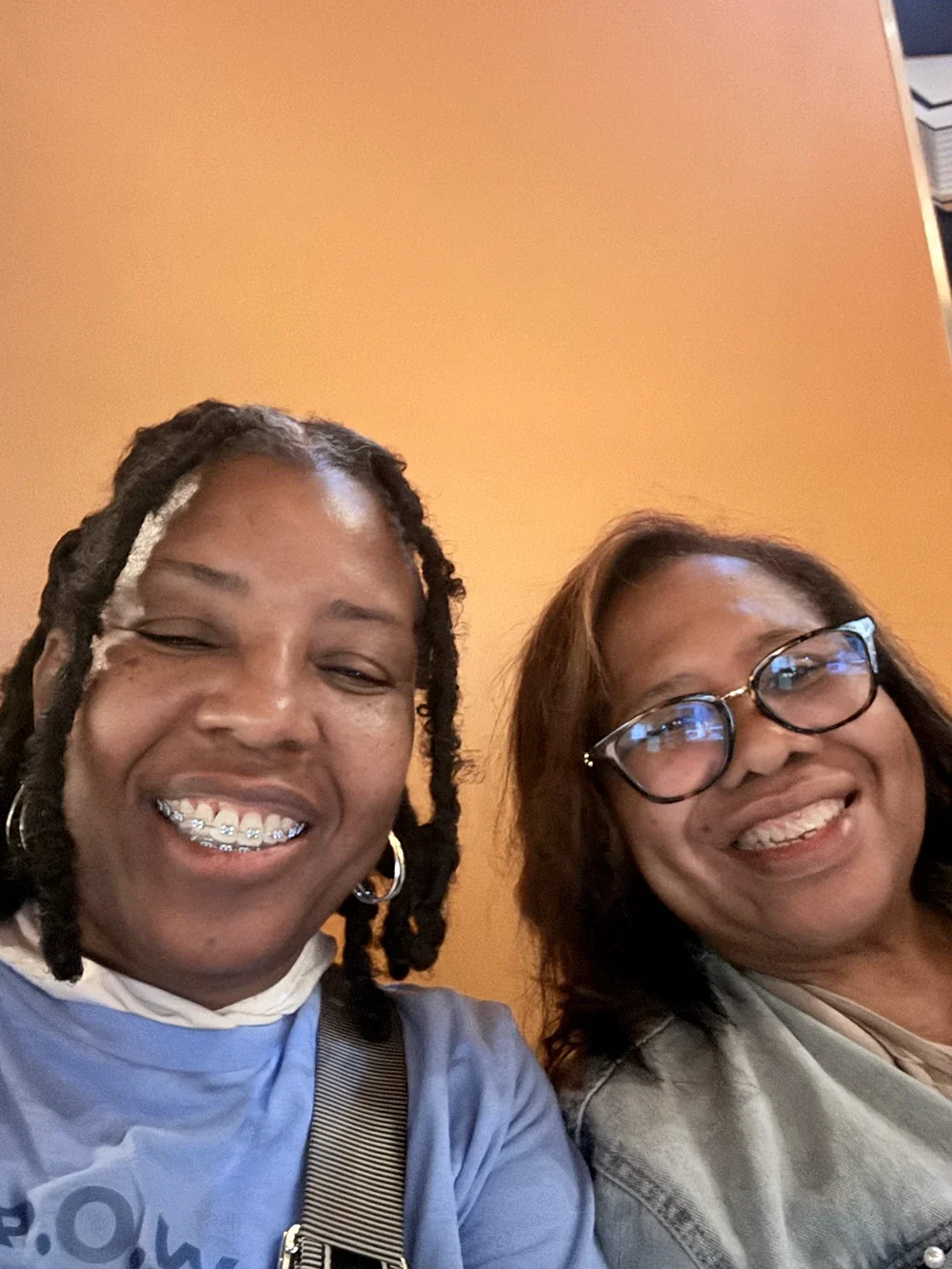 Two women smiling and taking a selfie indoors against an orange wall.