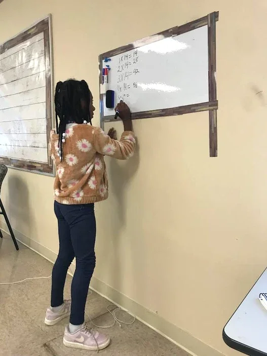 A young girl standing in front of a whiteboard on a beige wall, writing with a marker. She is wearing a brown sweater with daisy patterns, dark jeans, and light pink sneakers. The whiteboard has some math problems written on it, and there is a housep